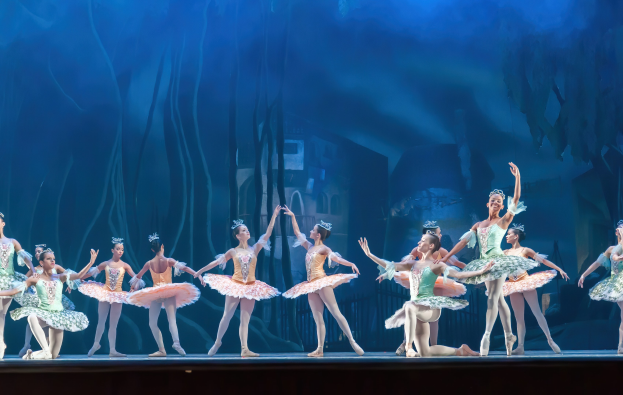 Eine Gruppe von Balletttänzerinnen in bunten Kostümen, die *Der Nussknacker* auf der Bühne der Royal Opera House tanzen, bewegen sich synchronisiert vor einem blauen Hintergrund.