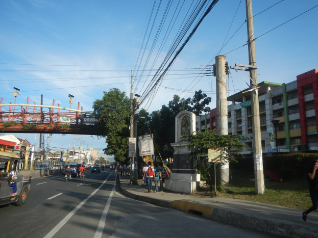 Eine belebte Stadtstraße mit Fahrzeugen, Fußgängern, Gebäuden, einer Brücke und Bäumen unter einem bewölkten Himmel.