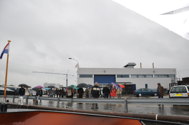 Eine Gruppe von Menschen im Freien, einige halten Schirme, mit einer Fahne an einem Mast, zwei Autos, ein Gebäude mit Fenstern, ein Turmkran, eine Straßenlaterne und eine Barrikade im Hintergrund.