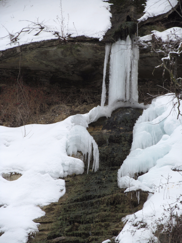 Ein kleiner Wasserfall ergießt sich über einen schneebedeckten, steinigen Hang in einem bewaldeten Gebiet, mit Eiszapfen an den Felsen und schneebedeckten Bäumen, die die Szene umgeben.