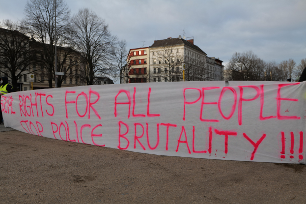 Gruppe von Menschen mit einer Fahne mit der Aufschrift "Rechte für alle Menschen Stoppt Polizeigewalt" vor einer Straßenlaterne, einem Schild, Bäumen, Gebäuden und einem bewölkten Himmel.