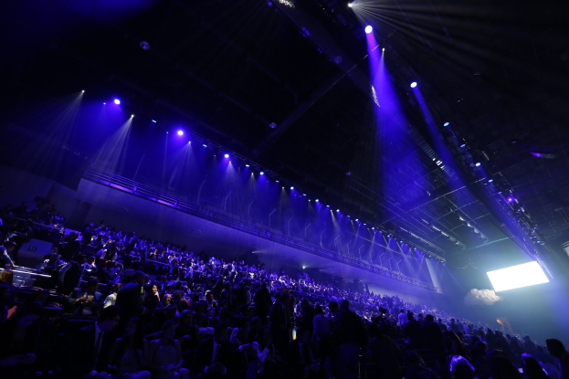 Eine große Menschenmenge sitzt in einem abgedunkelten Konzertsaal vor einer Bühne mit Deckenlichtern und einem Bildschirm auf der rechten Seite.