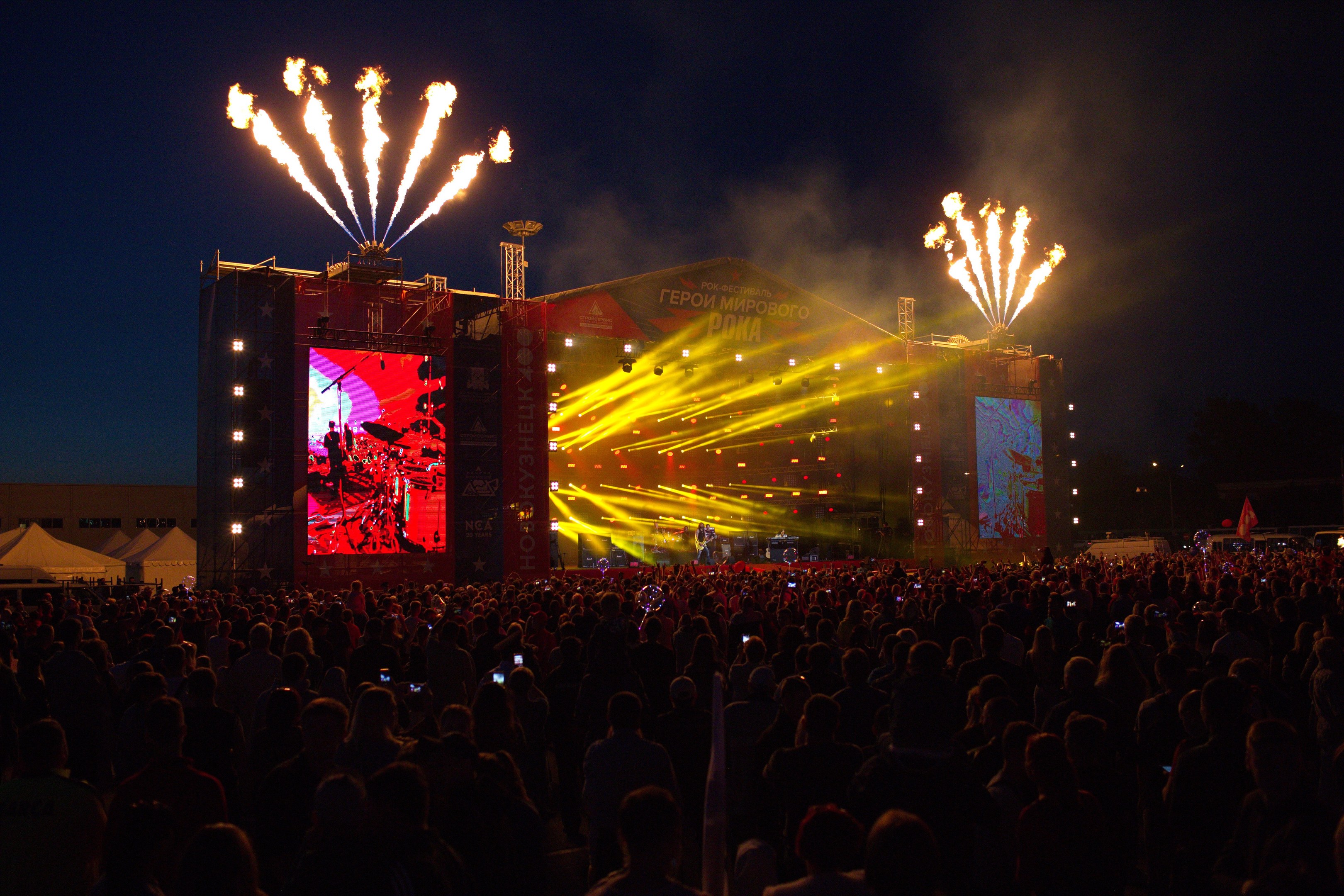 Eine große Menschenmenge bei einem Musikfestival schaut Performern auf einer hell erleuchteten Bühne zu, mit Zelten links und Feuerwerk am dunklen Himmel dahinter.