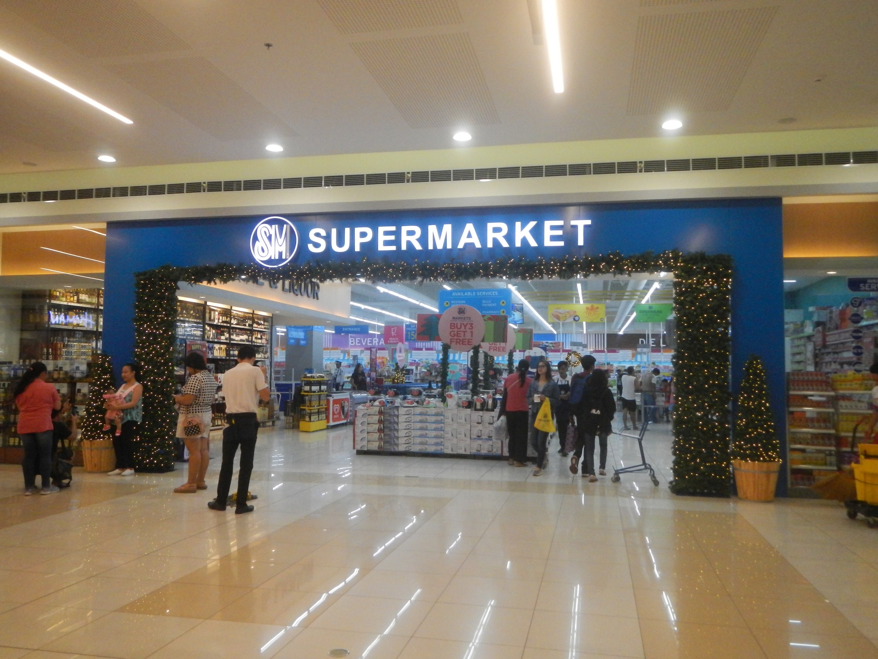 Eine Gruppe von Menschen vor einem Supermarkt, einige mit Einkaufstaschen, einem Einkaufswagen, einem Topf mit Pflanze, Regalen mit Gegenständen, Schildern und einer Decke mit Lampen an der Decke.