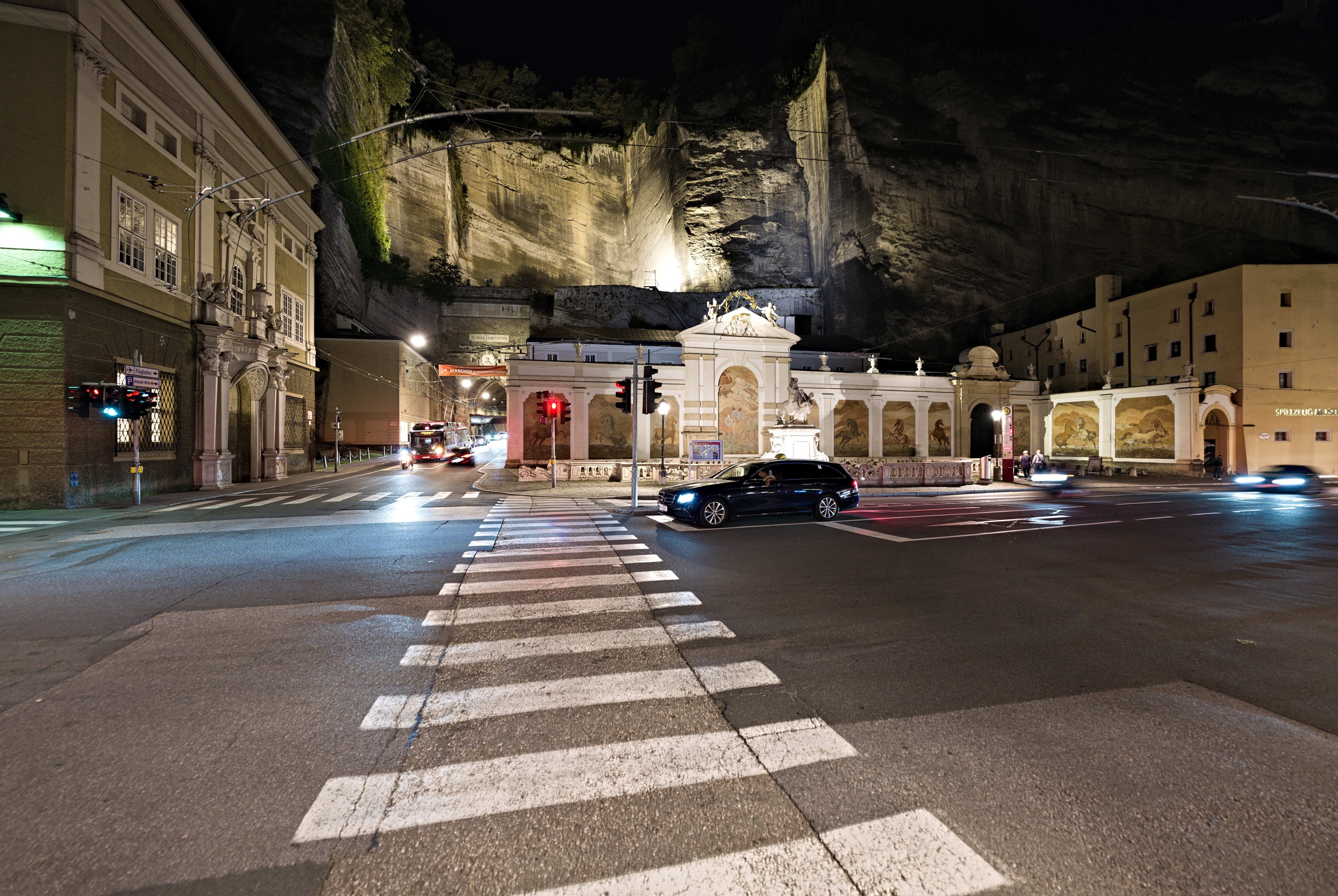 Eine nächtliche Straßenkreuzung mit fahrenden Autos, Straßenlaternen, Ampeln, beleuchteten Gebäuden, Laternenpfählen, einem Hügel im Hintergrund und einer Zebrastreifenmarkierung am Boden.