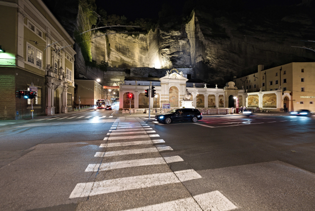 Eine nächtliche Straßenkreuzung mit fahrenden Autos, Straßenlaternen, Ampeln, beleuchteten Gebäuden, Laternenpfählen, einem Hügel im Hintergrund und einer Zebrastreifenmarkierung am Boden.