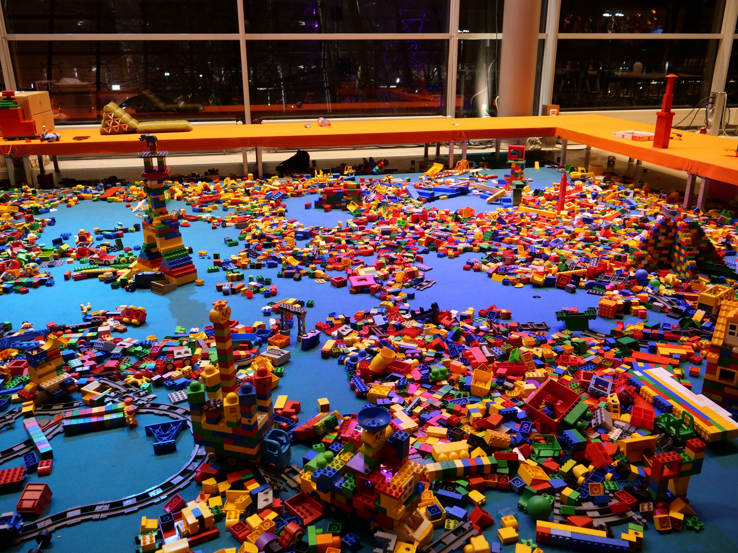 Ein großer Raum mit bunten Lego-Bauklötzen, die auf dem Boden verstreut sind, umgeben von Tischen und Säulen, und Glasfenstern im Hintergrund, die Menschen und Lichter zeigen.