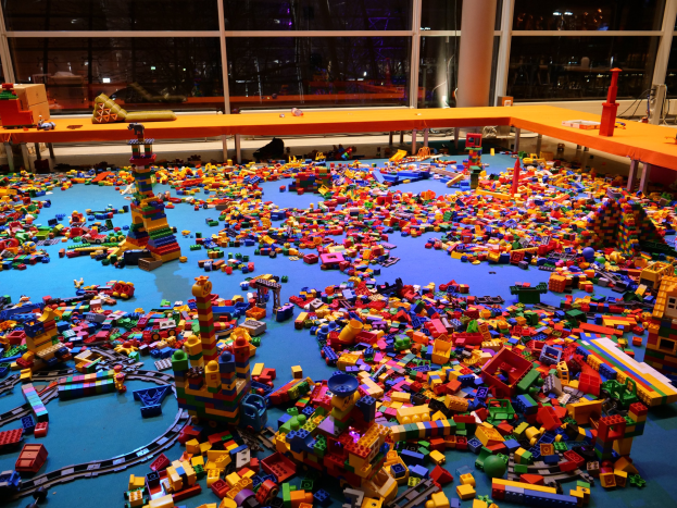 Ein großer Raum mit bunten Lego-Bauklötzen, die auf dem Boden verstreut sind, umgeben von Tischen und Säulen, und Glasfenstern im Hintergrund, die Menschen und Lichter zeigen.