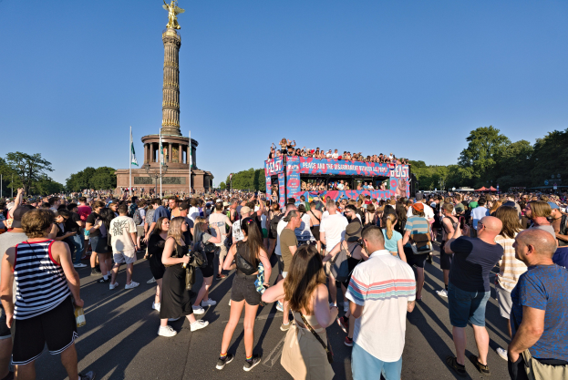 Große Menschenmenge versammelt sich vor einem Denkmal in Berlin, Deutschland, mit Fahnen, einem Gebäude mit Säulen und einer Statue, Bäumen und einem klaren blauen Himmel.