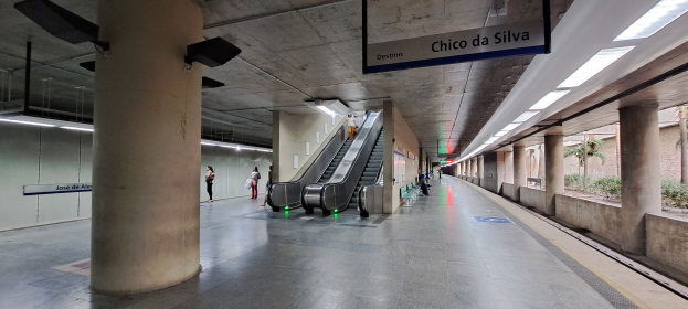 Eine U-Bahn-Station mit einer Rolltreppe in der Mitte, Menschen, die herumgehen, Säulen, Namensschilder, Deckenleuchten und Pflanzen und Bäume auf der rechten Seite.