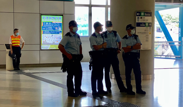 Eine Gruppe von Polizisten in Masken steht vor einem Gebäude mit Glastüren, Schildern mit Aufschriften, einer Säule und einem Geländer, mit Bäumen und Fahrzeugen im Hintergrund.