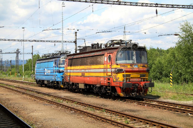 Ein Zugtriebwagen auf Schienen mit elektrischen Masten, Kabeln, Bäumen, Pflanzen und Wolken am Himmel.