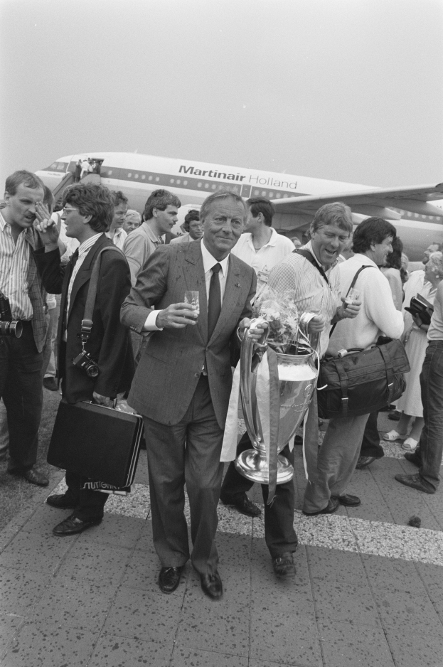 Ein Mann im Anzug und Krawatte hält eine Trophäe vor einem Flugzeug, umgeben von einer Gruppe von Menschen, einige halten Taschen und einer hält eine Kamera, auf einem Schwarz-Weiß-Bild.