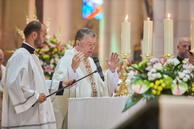 Der Bischof der Kirche des Heiligen Grabes steht an einem Rednerpult mit einem Mikrofon und spricht zu einer Gruppe von Menschen. Rechts von ihm befindet sich ein Tisch mit Kerzen und Blumen, und im Hintergrund sind Blumensträuße und ein Buntglasfenster zu sehen.