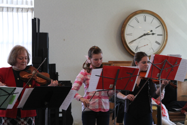 Drei Frauen stehen auf der Bühne und spielen Geigen an Musikständern, während ein sitzendes Publikum zuhört; eine Uhr ist an der weißen Wand im Hintergrund angebracht, und ein weißer Vorhang ist links im Bild zu sehen.