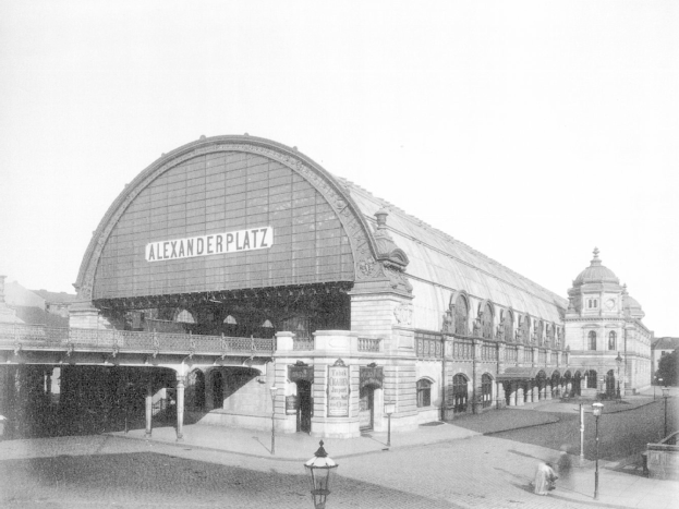 Ein Schwarz-Weiß-Foto von Alexanderplatz in Berlin, Deutschland, mit einem großen Gebäude mit Säulen, Bögen und einem Namensschild, Laternen, Menschen auf der Straße, Bäumen und einem klaren Himmel im Hintergrund.
