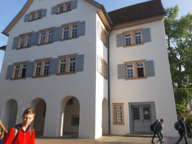 Eine Gruppe von Menschen, einige mit Taschen, geht vor einem großen weißen Gebäude mit Fenstern und Türen vorbei, umgeben von Bäumen unter einem klaren blauen Himmel.