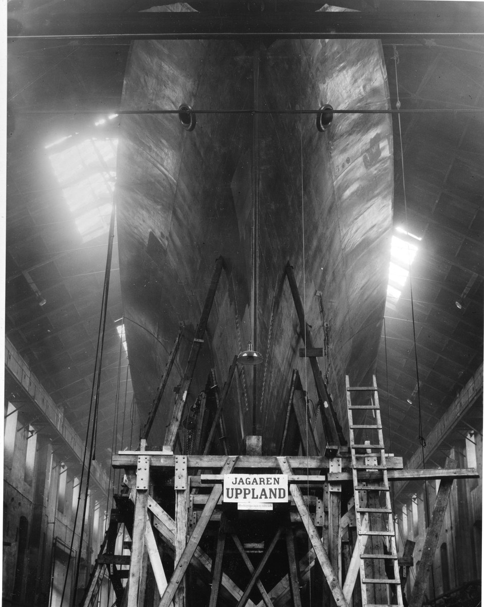 Schwarzes und weißes Foto eines großen Boots in einem Schiffbau, das eine Leiter, hölzerne Pfosten, eine Schautafel, Metallrahmen, Deckenleuchten und Fenster zeigt.