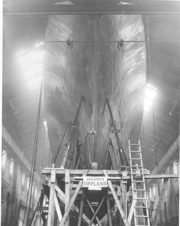 Schwarzes und weißes Foto eines großen Boots in einem Schiffbau, das eine Leiter, hölzerne Pfosten, eine Schautafel, Metallrahmen, Deckenleuchten und Fenster zeigt.