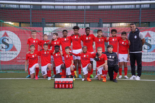 Eine Gruppe junger Männer in roten T-Shirts und schwarzen Shorts steht auf einem Fußballfeld vor einer Kiste roter Gegenstände, im Hintergrund sind Banner mit 'FC Red Black Eglaute - Champions League' zu sehen.