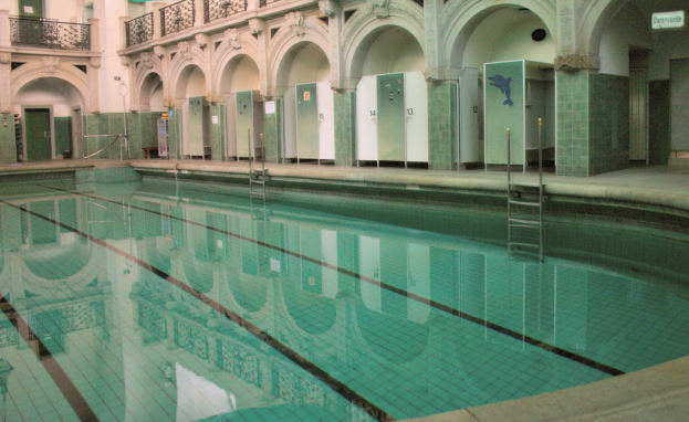 Hallenbad mit klarem Wasser, umgeben von gefliestem Boden, Bogengängen, Säulen, Geländern, Türen und Texttafeln in einem großeen Gebäude.