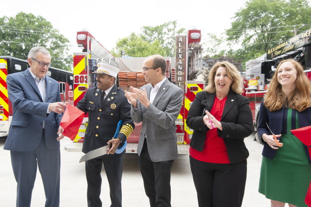 Eine Gruppe von Menschen steht vor einem Feuerwehrauto, klatscht und lacht, mit zwei Personen, die eine Schere und ein rotes Tuch halten, bei einer Eröffnungszeremonie der Feuerwehr Alexandria, mit Bäumen, Drähten und einem klaren blauen Himmel im Hintergrund.
