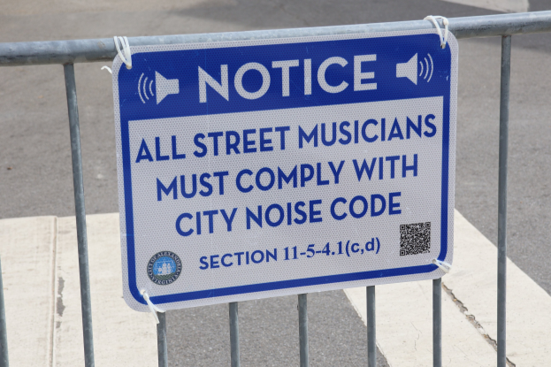 Hinweistafel an einem Zaun mit einem Schild, auf dem "Hinweis: Alle Straßenmusiker müssen die Stadtlärmvorschriften einhalten" steht, und einer Straße im Hintergrund.