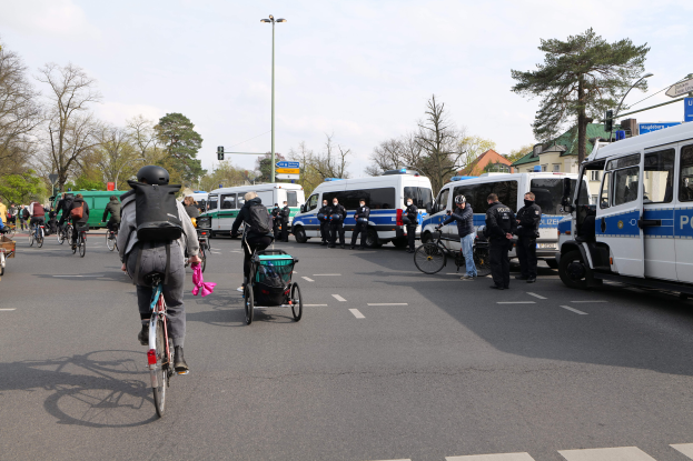 Eine Gruppe von Menschen, die auf Fahrrädern eine von Polizeiwagen und anderen Fahrzeugen gesäumte Straße entlangfahren, einige tragen Helme und Taschen, andere schieben Kinderwagen, mit Bäumen, Laternen, Verkehrsampeln, Schildern, Gebäuden und einem bewölkten Himmel im Hintergrund.