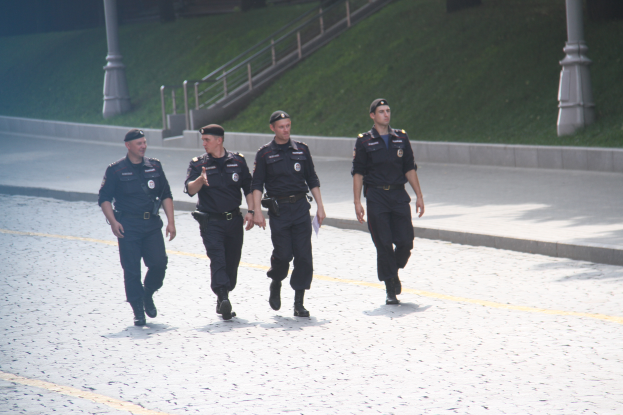 Drei Polizisten in Uniform gehen eine Straße entlang mit Pfosten, Geländern, Treppen und Gras im Hintergrund.