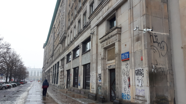 Eine Person geht auf einem Gehweg an einem Gebäude mit Graffiti vorbei, mit parkenden Fahrzeugen, Bäumen und Gebäuden im Hintergrund unter einem Himmel.