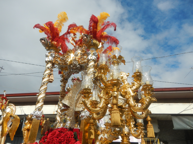 Ein großer goldener und roter Festwagen, geschmückt mit Blumen und anderen Dekorationen bei einer Karnevalsumzug, mit einem Gebäude, Strommasten mit Drähten und einem bewölkten Himmel im Hintergrund.