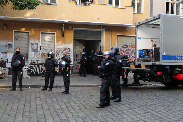 Gruppe von Polizisten in Helmen und taktischer Ausrüstung mit Waffen, die vor einem Gebäude mit Graffiti an den Wänden, Fenstern und Rollläden stehen, mit einem Lastwagen und Bäumen im Hintergrund.