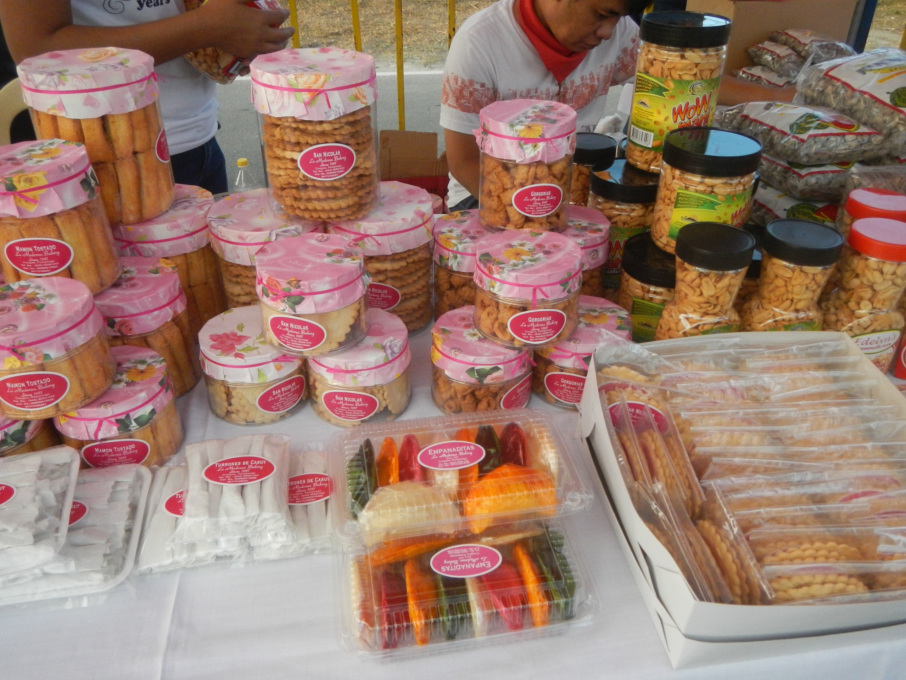 Eine Gruppe von Menschen steht um einen Tisch mit verschiedenen Lebensmitteln auf einem Food-Festival, im Hintergrund ein Gel├Ąnder und eine Stra├če.