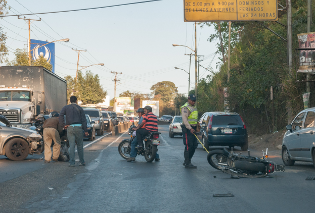 Eine Gruppe von Menschen steht um ein verunglücktes Motorrad auf der Seite einer Straße mit mehreren Fahrzeugen, darunter ein Lastwagen, und Hintergrundelementen wie Bäumen, Pfählen, Lampen, Schildern und dem Himmel.