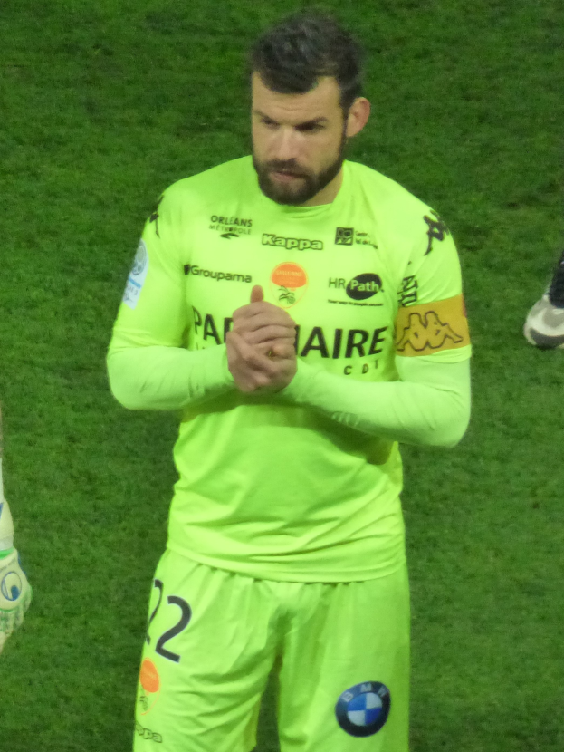Ein Mann in einem gelben Torwarttrikot, grüner T-Shirt und Shorts steht auf einem Fussballfeld und klatscht in die Hände, mit einem anderen Bein auf der linken Seite des Bildes sichtbar.
