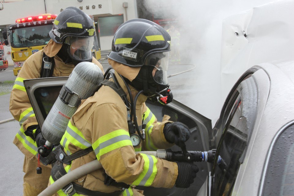 Zwei Feuerwehrleute in Schutzausrüstung verwenden einen Schlauch, um ein brennendes Auto zu löschen, mit Rauch und Fahrzeugen und einem Gebäude im Hintergrund.
