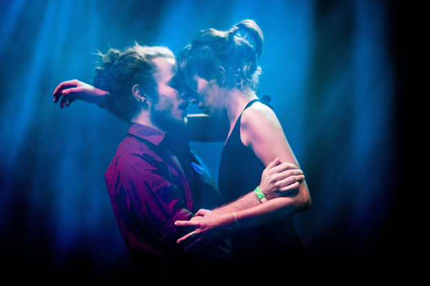 Zwei Menschen umarmen und küssen sich auf einer Bühne mit abgedunkeltem Hintergrund.