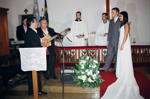 Eine HochzeitsSzene in einem Raum mit zwei Pastoren, einer Braut, Bräutigam und drei Musikern; ein Podium mit Stoff vor den Musikern; ein Tisch mit Kerzen hinter den Pastoren; ein Kerzenhalter hinter dem Paar; ein Bukett, Lautsprecher, Taschen und roter Teppich auf dem Boden; sowie Flaggen, eine Wand mit einem Kreuz, einen Schrank und einen Stuhl im Hintergrund.