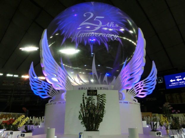 Ein großer Kristallball mit Flügeln oben drauf, umgeben von Blumenbouquets, Namensschildern und anderen Gegenständen auf dem Boden, mit Tafeln, Lichtern und einem Bildschirm im Hintergrund bei einer 25-jährigen Jubiläumsfeier.
