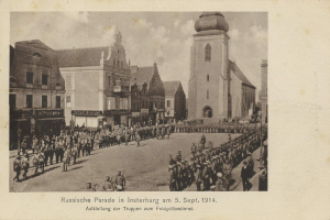 Schwarzes und weißes Foto einer Parade in Insterburg 1914 mit marschierenden und stehenden Menschen, Gebäuden im Hintergrund und Wolken am Himmel.