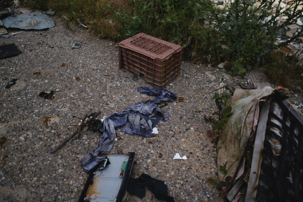 Ein Haufen Müll einschließlich eines Plastikbehälters, Kleidung und anderer Gegenstände, die auf dem Boden neben einem Zaun verstreut sind, mit Pflanzen und Gras im Hintergrund.