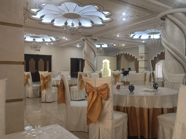 Großer Raum mit Tischen und Stühlen für eine Hochzeitsfeier eingerichtet, Stühle mit Tüchern drapiert und dekoriert, Wände mit Türen und Vorhängen versehen, Decke mit Lichtern und Kronleuchtern geschmückt.