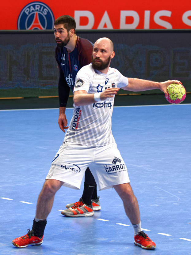 Zwei Männer spielen Handball auf einem Platz, einer hält den Ball, mit einem Schild im Hintergrund, auf dem 'Paris Saint-Germain vs Paris Saint Germain' steht.