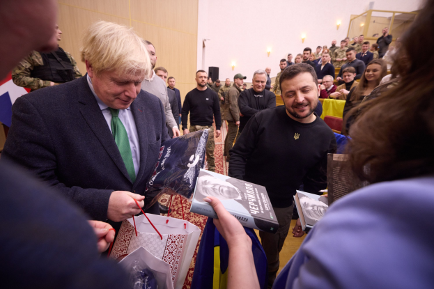 Ein Mann im Anzug und Krawatte unterschreibt Autogramme für eine Menschenmenge, einige halten Taschen und Bücher, mit einer beleuchteten Wand im Hintergrund.