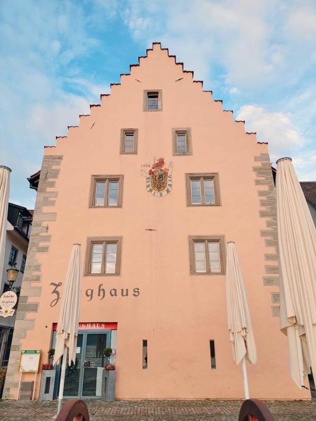 Pinkes Gebäude mit einem zentralen Uhrenturm, umgeben von Fenstern, Schirmen, einem Schild, einer Straßenlaterne, Topfpflanzen und einem bewölkten Himmel, repräsentiert München, Deutschland.