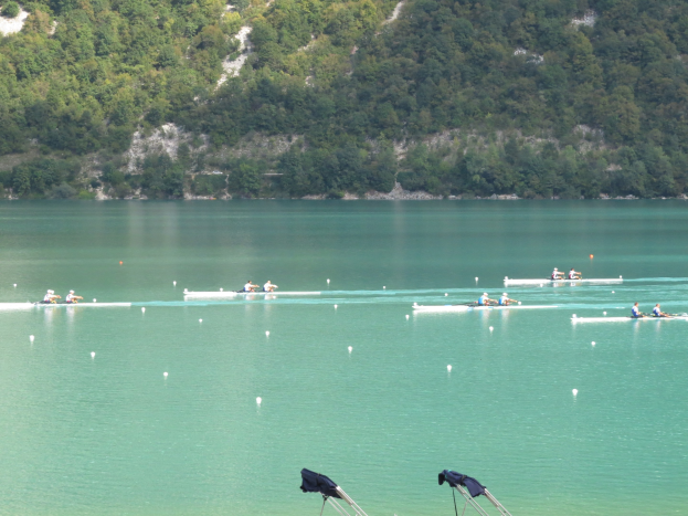 Gruppe von Menschen beim Ruderwettbewerb auf einem Gewässer mit Bäumen und einem Hügel im Hintergrund, Kleidung auf Ständen am Wasserrand.