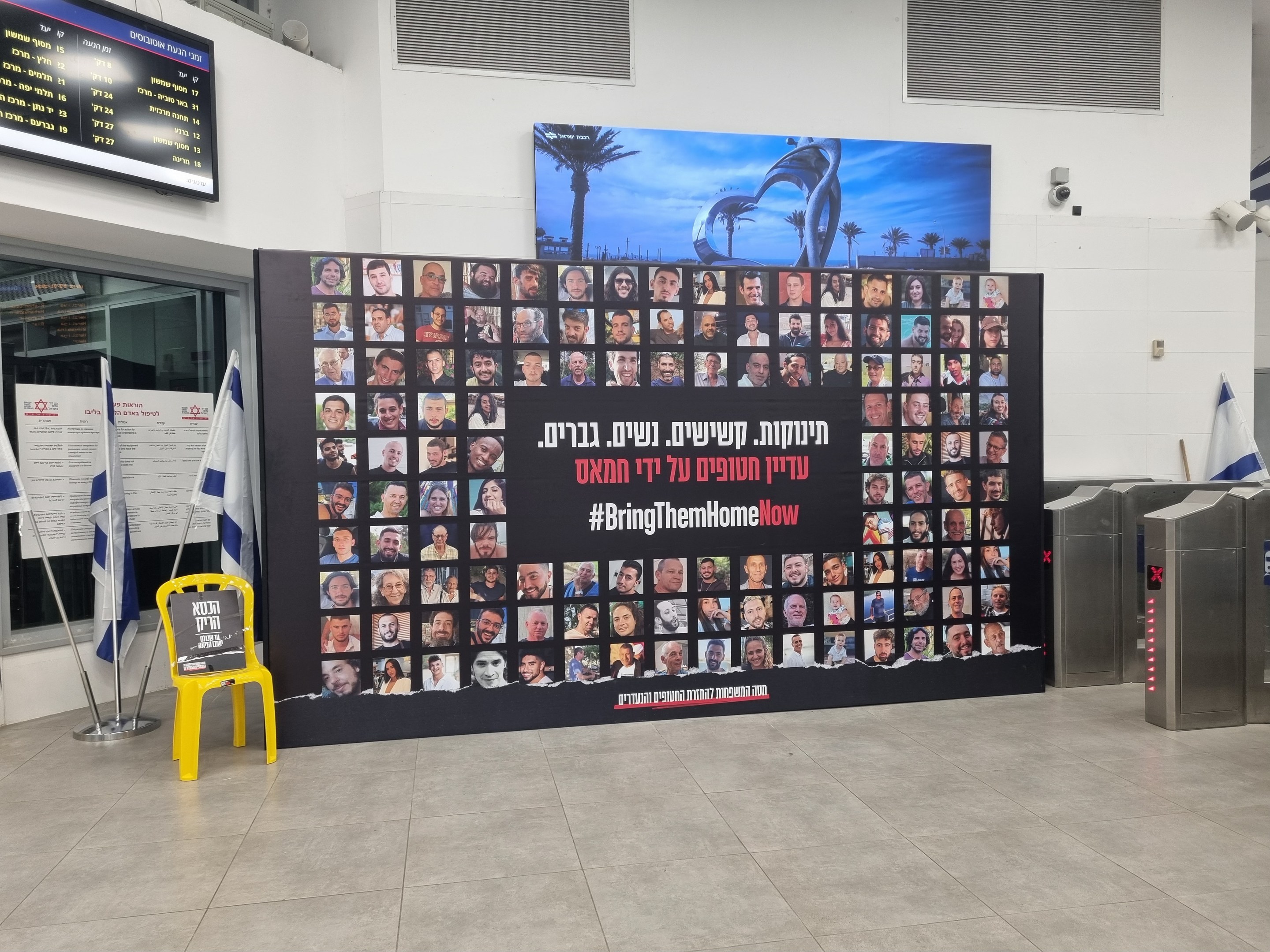 Eine große Wand zeigt verschiedene Bilder, einen gelben Stuhl, israelische Regierungsflaggen auf Ständern, eine Tafel mit der Aufschrift "Bring Them Home Now", einen montierten Bildschirm, Deckenleuchten und andere Gegenstände.