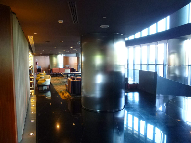 Eine geräumige First-Class-Lounge am Hong Kong International Airport mit großen Fenstern, hohen Decken mit elektrischen Leuchten, Sitzmöglichkeiten einschließlich Stühlen und Sofas, Tischen, einem Teppichboden und einer zentralen Säule.