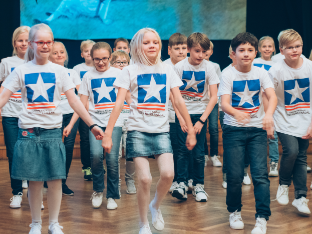 Eine Gruppe von Kindern in weißen Hemden und blauen Shorts tanzt auf einem Holzboden vor einer Wand mit einem Bildschirm und nimmt an einem kunstnädler Tanzwettbewerb teil.