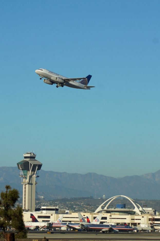 Eine Flughafenansicht mit einem weißen Kontrollturm, parkenden Flugzeugen, fernen Gebäuden und einem Flugzeug am Himmel.